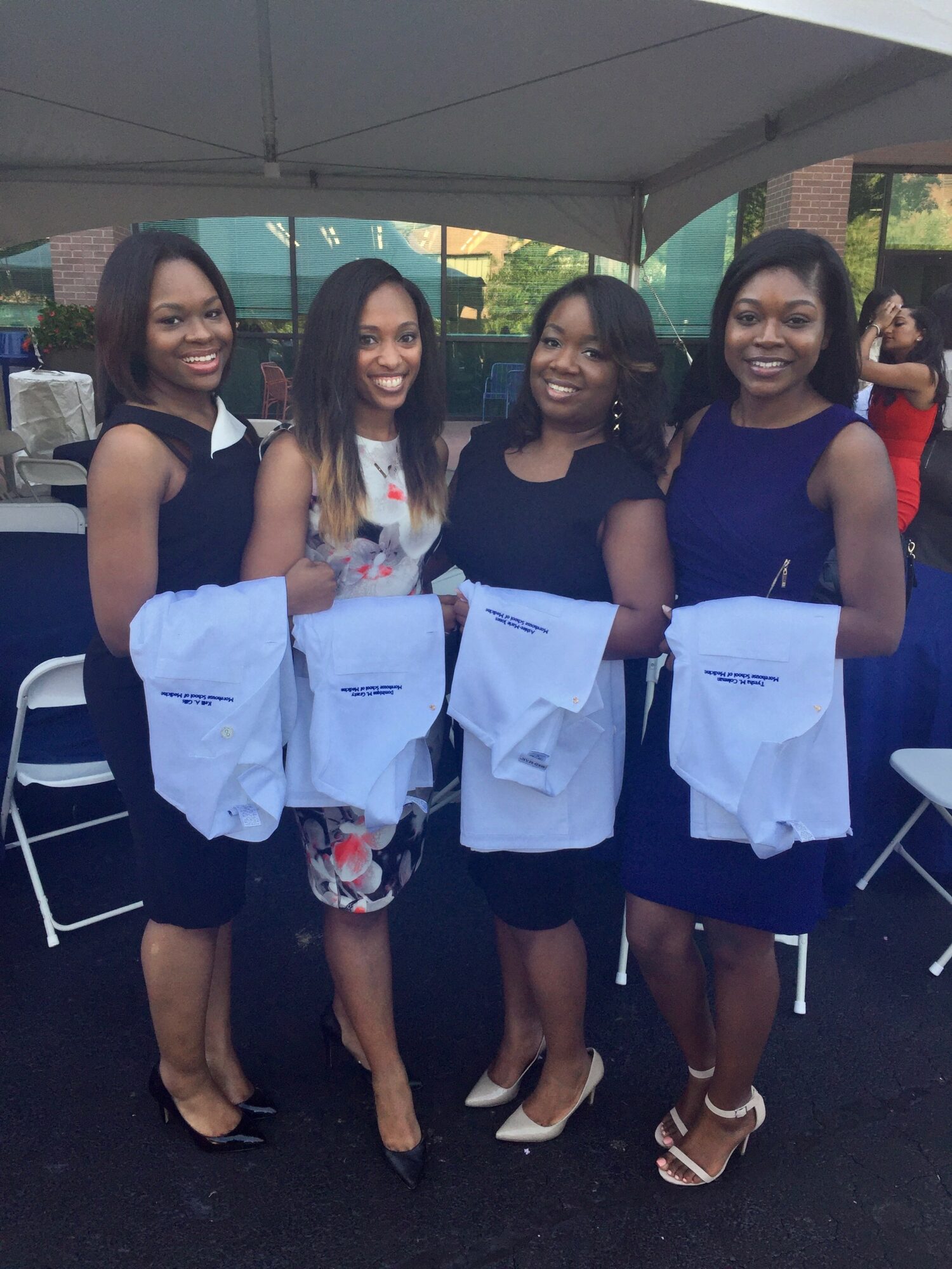 Four women standing outdoors under a tent, holding white cloths, smiling, with chairs and trees in the background.