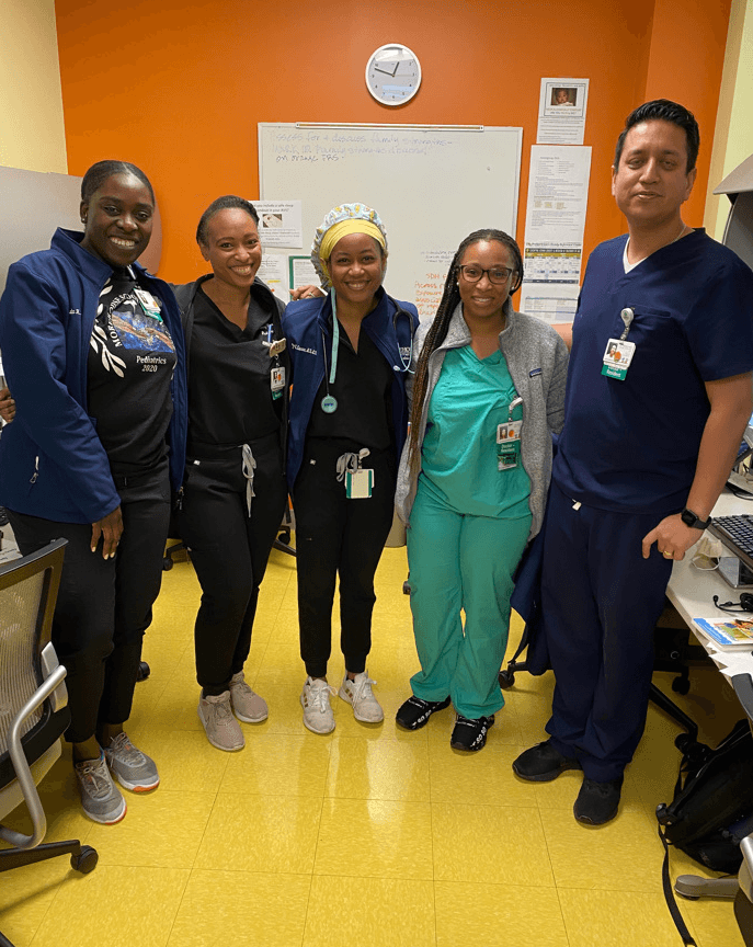 Group of five healthcare professionals standing in a room with orange walls and a whiteboard, smiling at the camera.