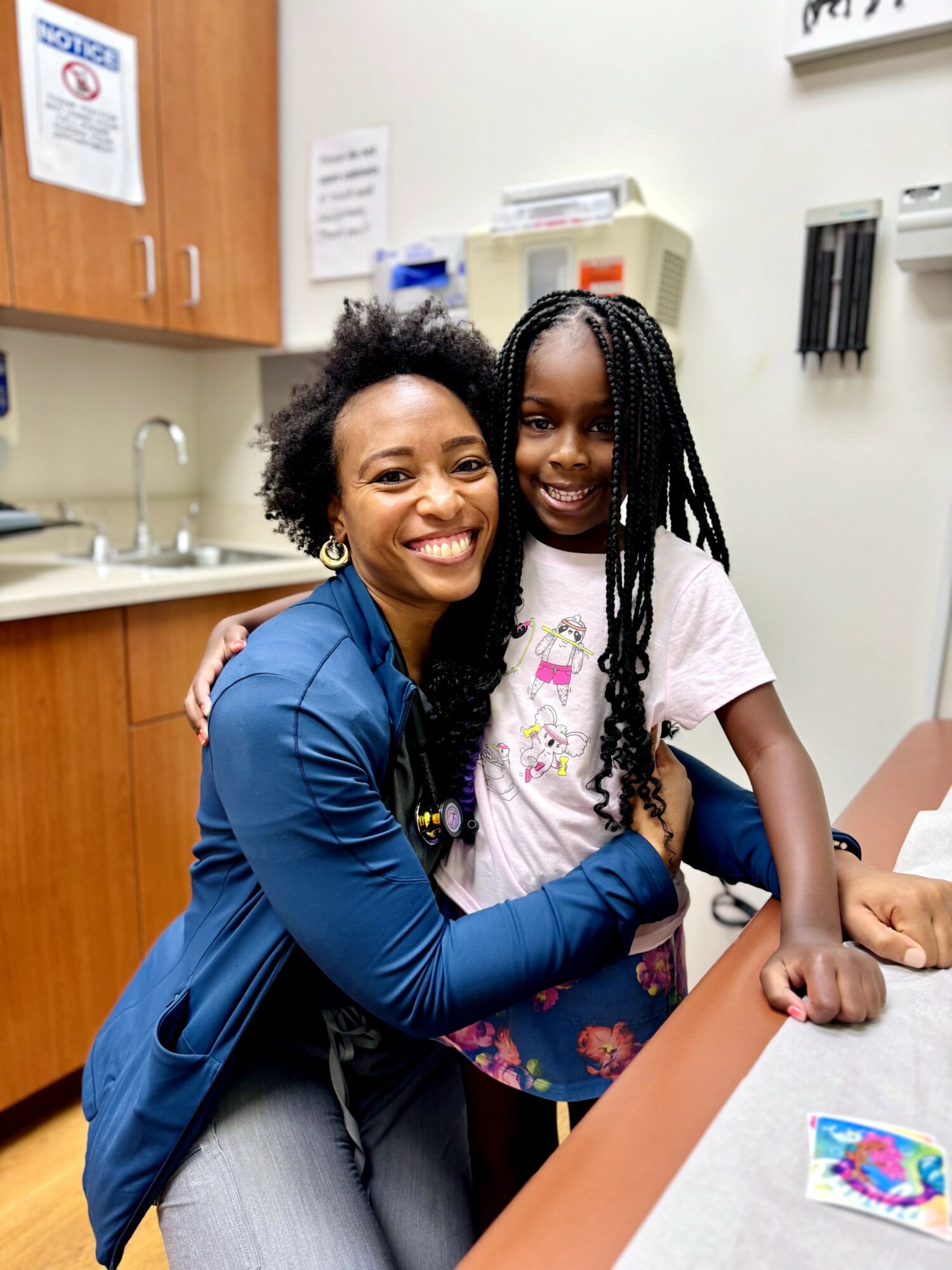 Woman and girl smiling, hugging in a room with cabinets and medical equipment, woman wearing a blue jacket, girl in a pink shirt.