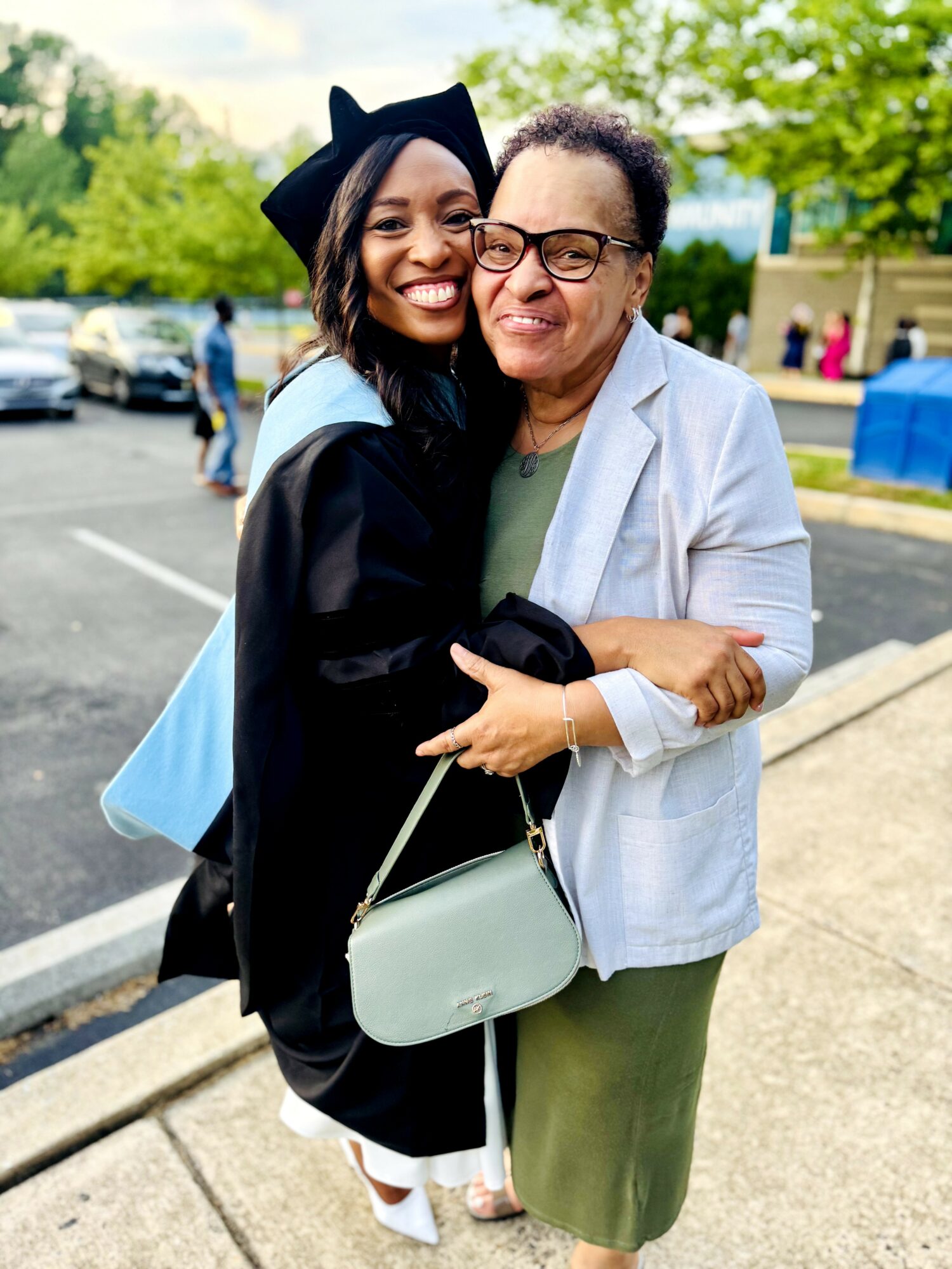 Graduate in cap and gown hugging an older woman outdoors, smiling, with trees and parked cars in background.