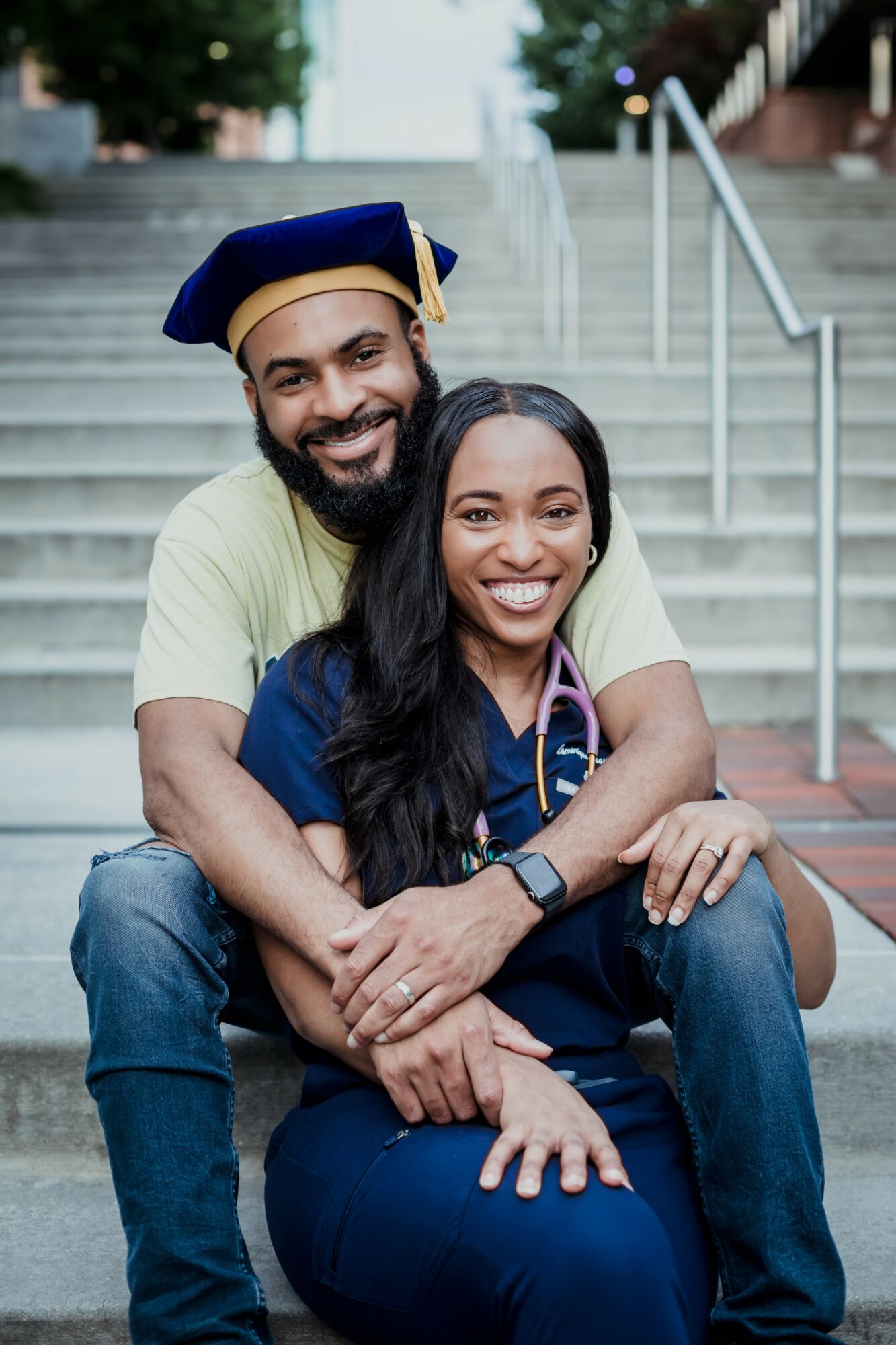 A smiling man and woman sit on stairs outdoors, with the man embracing the woman from behind, both wearing casual clothes.