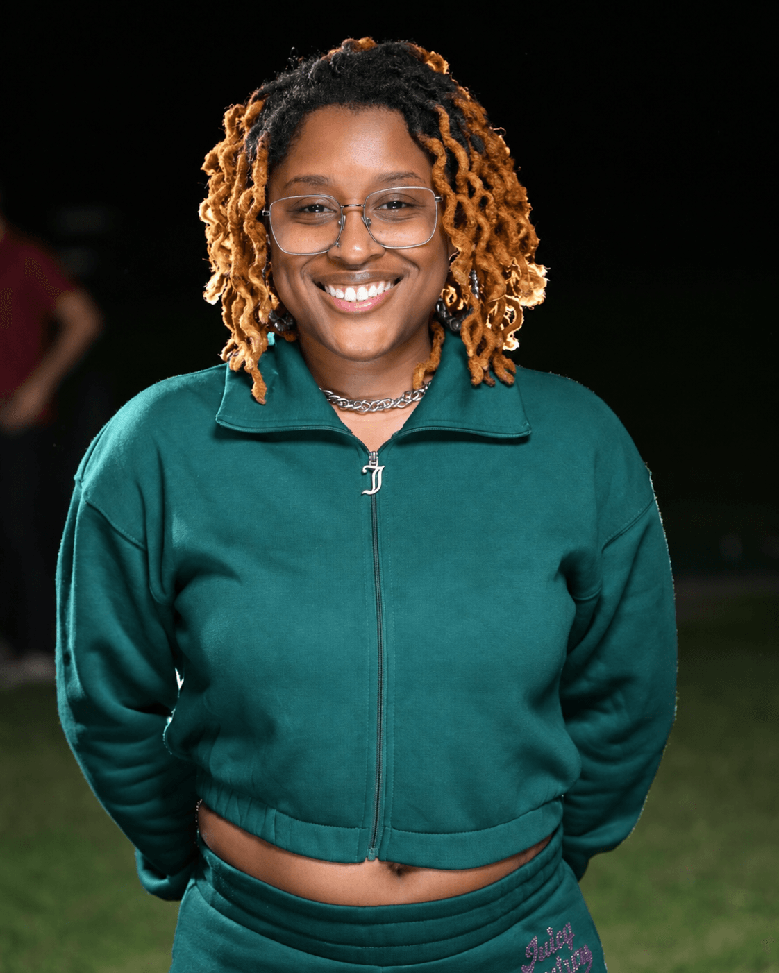 Smiling woman with glasses and curly, shoulder-length hair wearing a green zip-up jacket outdoors at night.