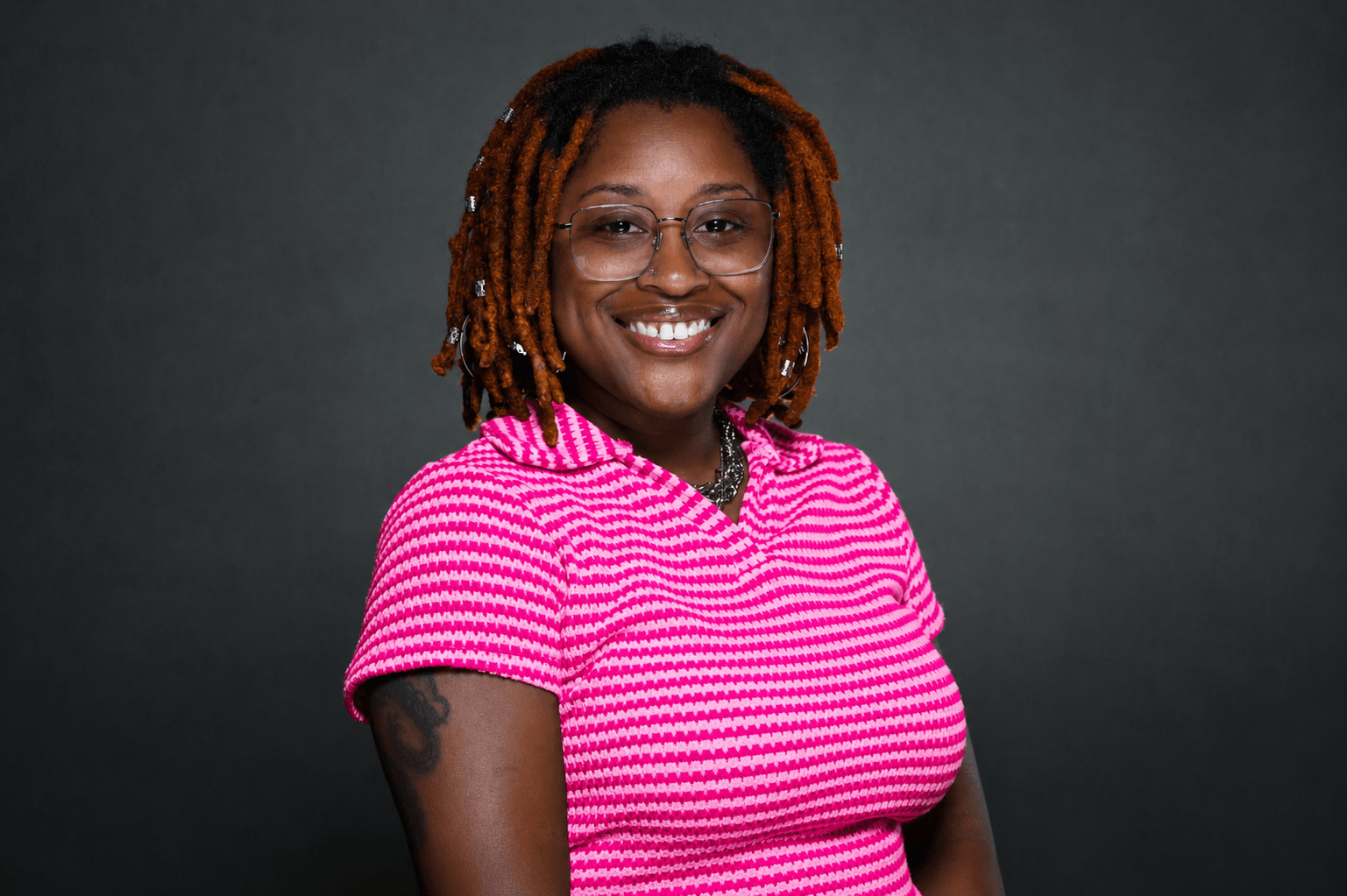 Smiling woman with glasses and dreadlocks wearing a pink and white striped shirt against a dark background.