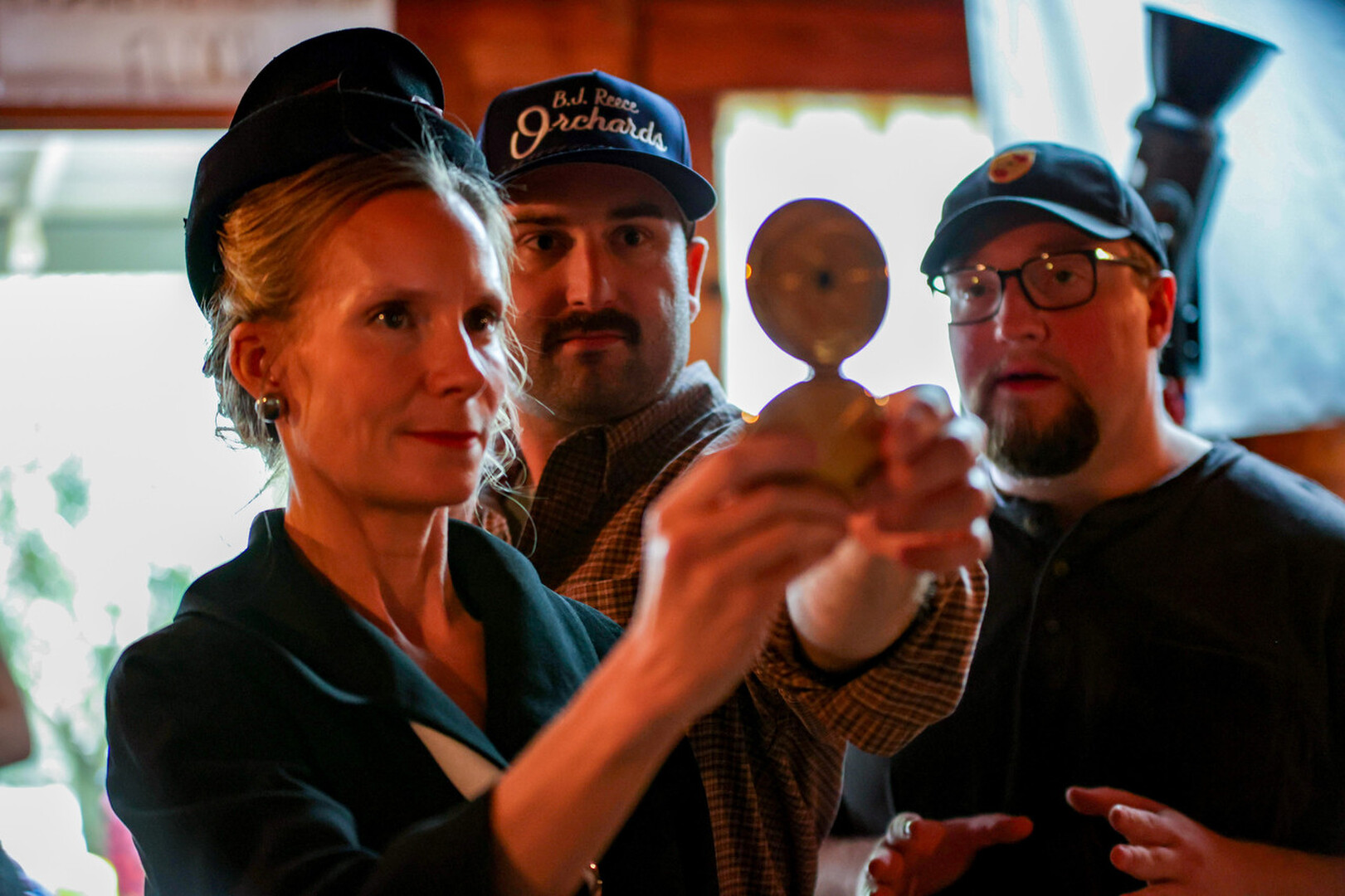 Three people indoors, woman holding a small object, two men watching, background with window and light, casual setting.