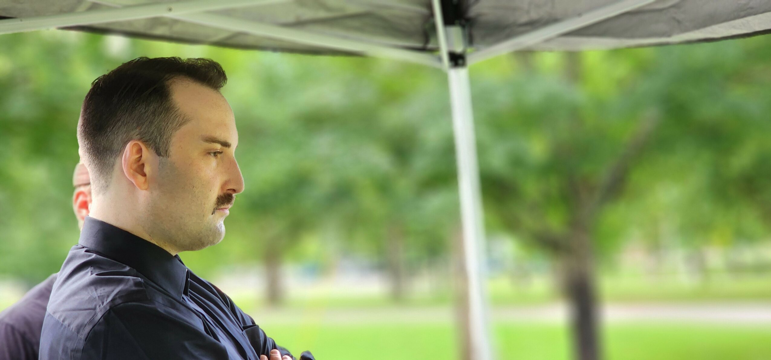 Man with closed eyes standing outdoors under a canopy, with trees and greenery in the background.
