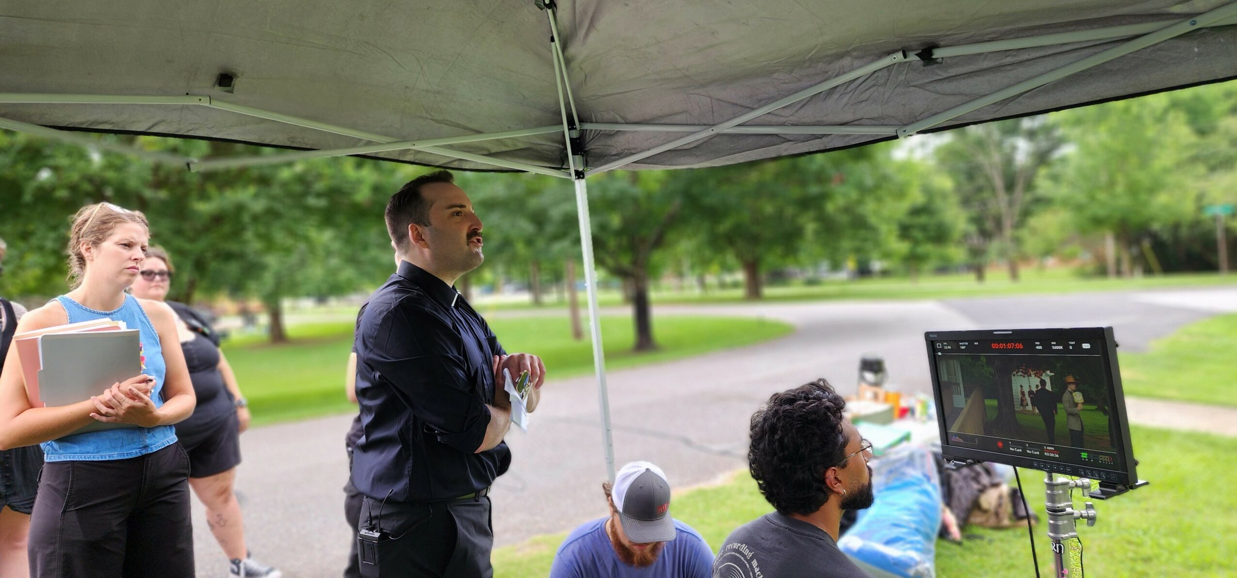 Group of people gathered outdoors under a canopy, with a man speaking and a monitor displaying a presentation.
