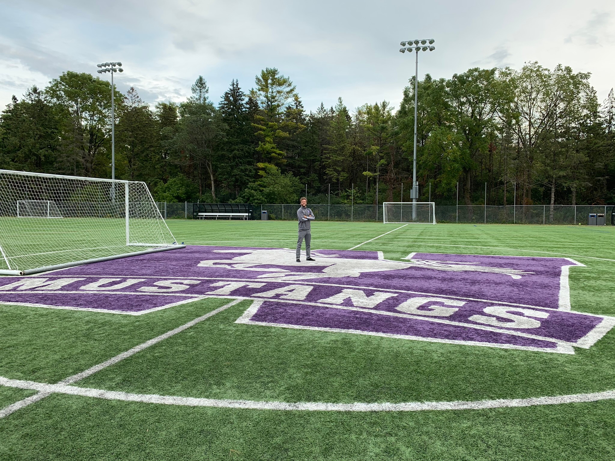 Person standing on a sports field with purple and white markings, surrounded by trees and light poles.