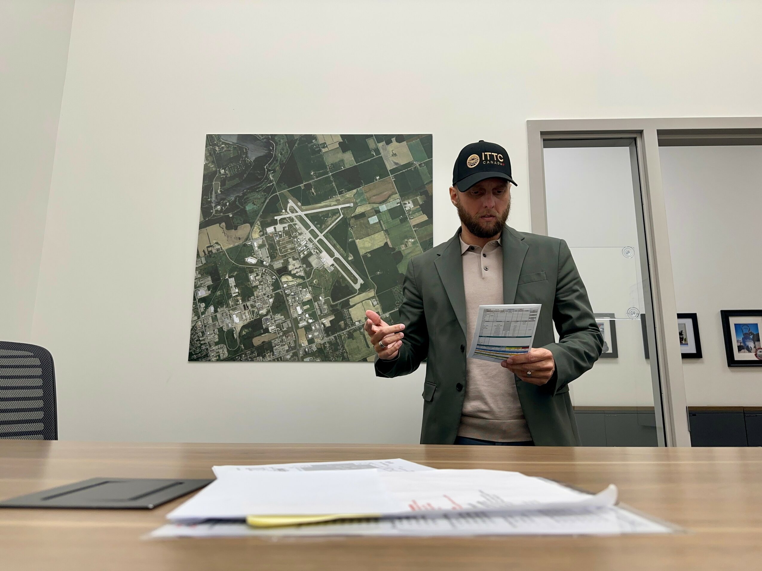 Man in suit and cap standing at table, holding papers, in office with large map on wall behind him.
