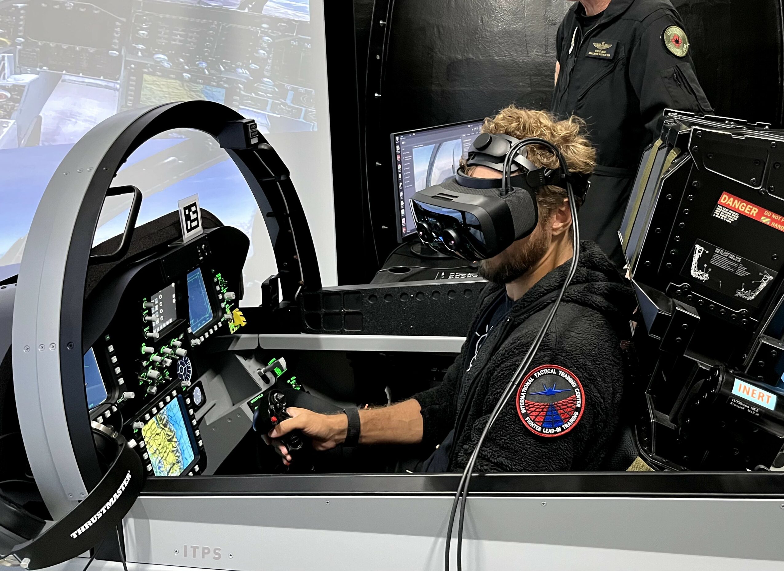 Person wearing a virtual reality headset and seated at a control panel with multiple screens and controls.