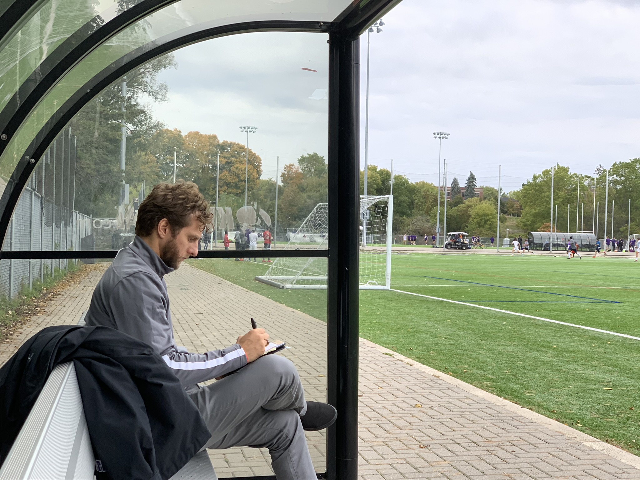 Man sitting on a bench inside a glass shelter, writing in a notebook, with a sports field outside.