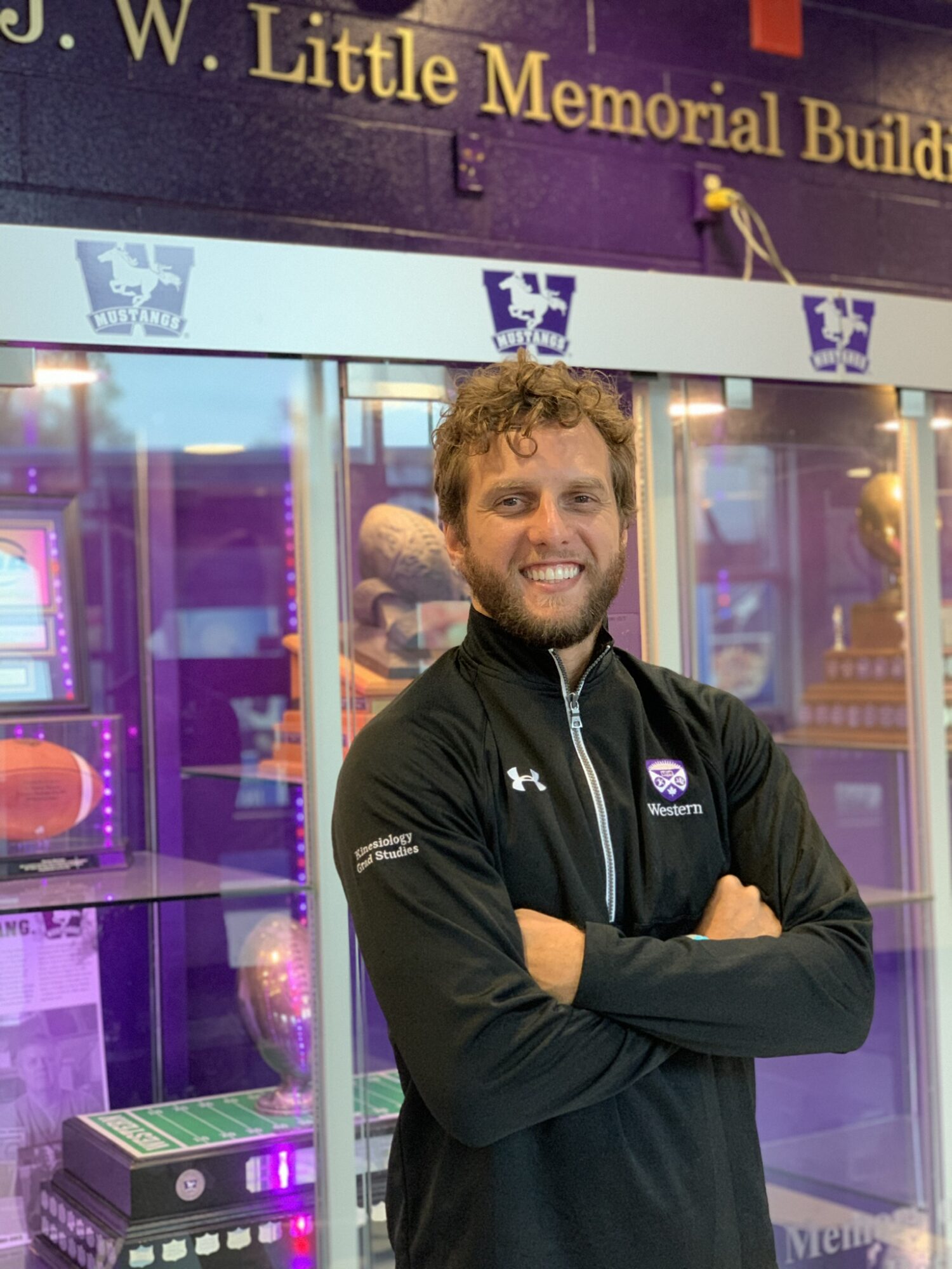 Man with curly hair and beard smiling, arms crossed, standing in front of colorful display case, wearing a black jacket.