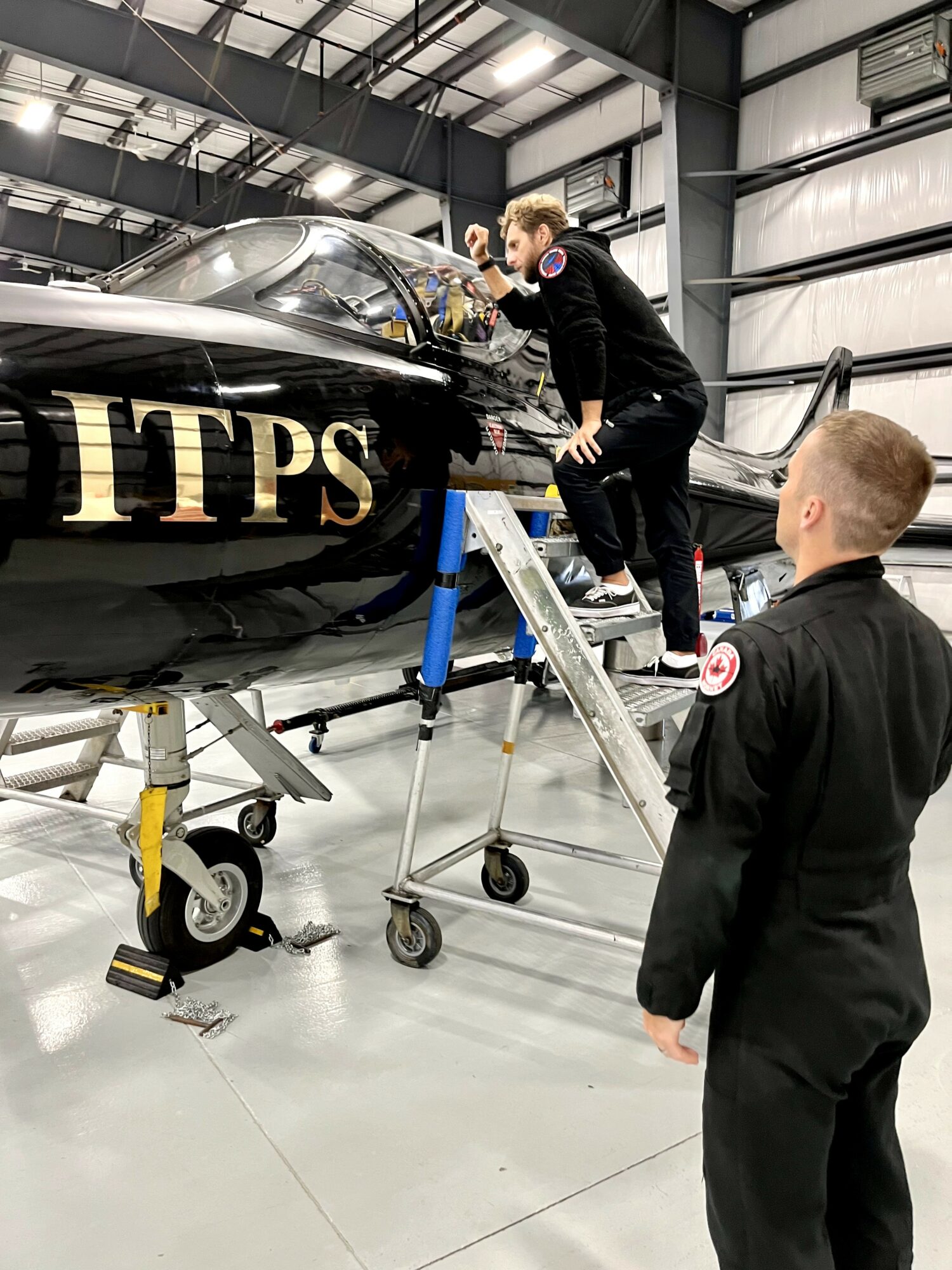 Two people in black uniforms inspect a jet aircraft inside a hangar, one on a ladder and the other on the ground.