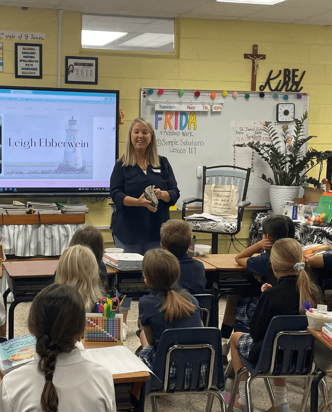 Woman standing in front of a classroom with students seated at desks, a whiteboard, and a large plant, teaching a lesson.
