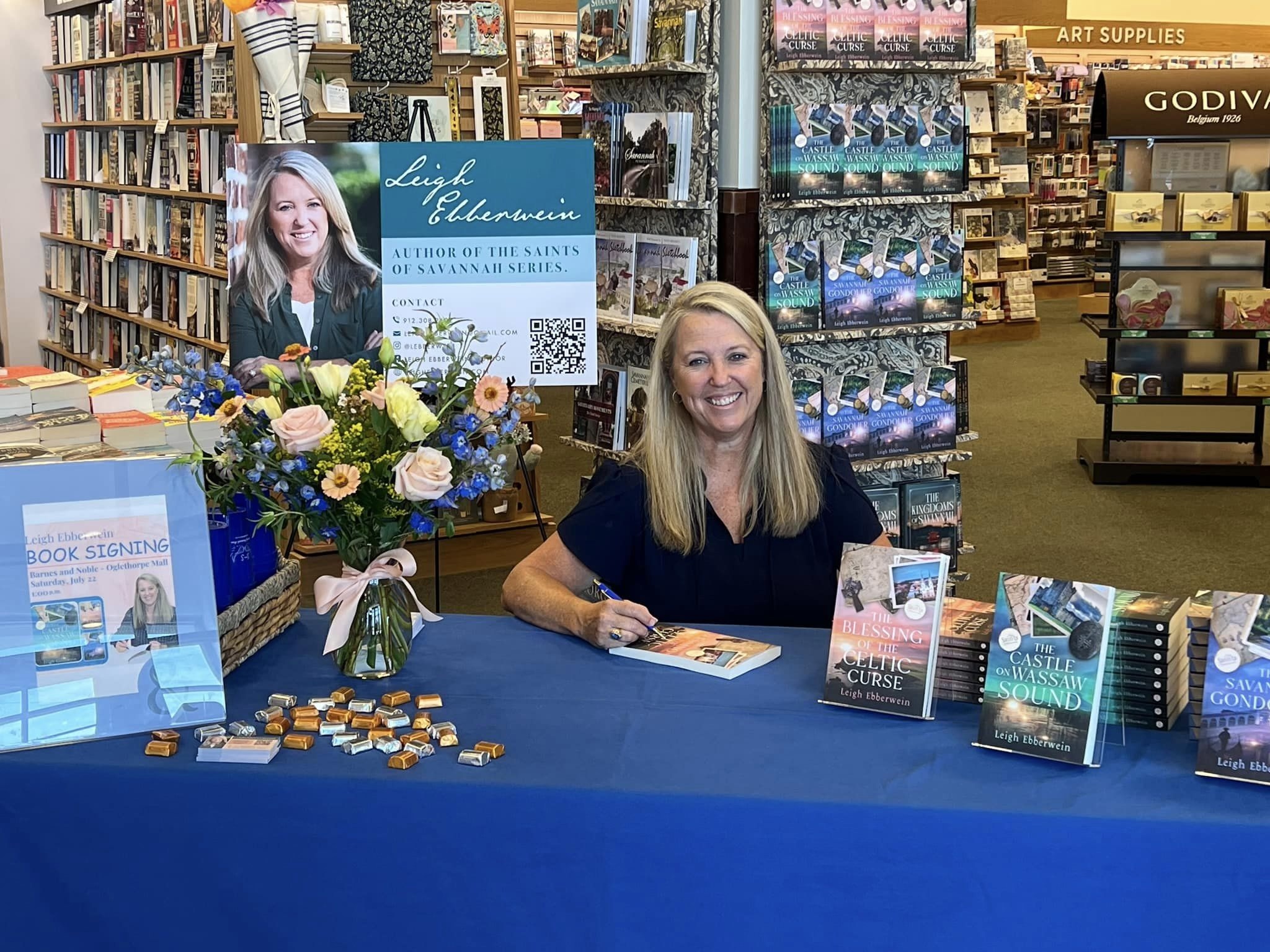 Woman sitting at a table with books and a flower arrangement inside a bookstore, smiling at the camera.