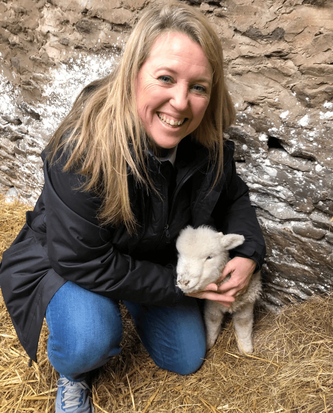 Woman crouching outdoors holding a small white lamb, smiling, with rocky background and dry grass.