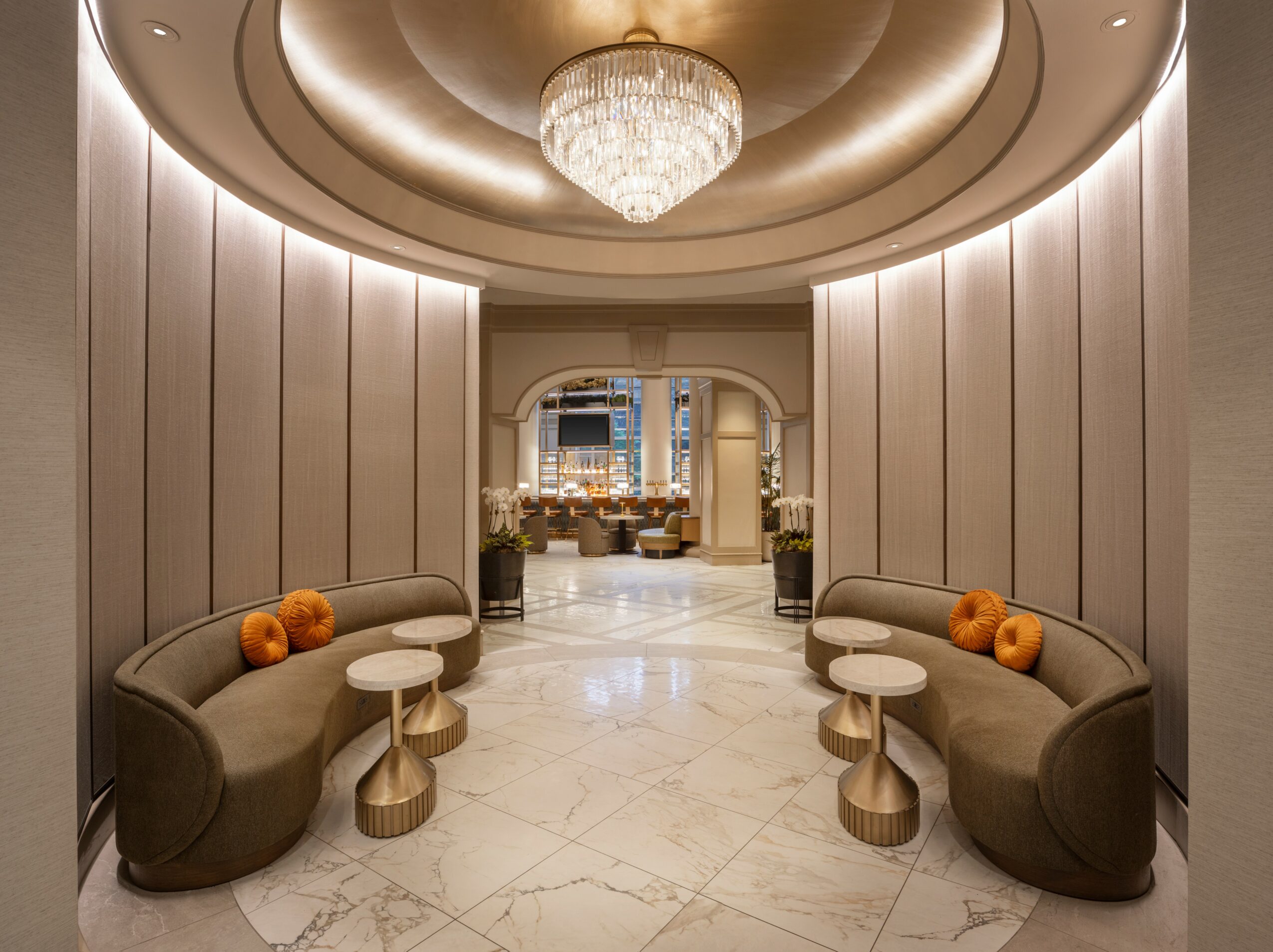 Elegant lobby with curved sofas, small tables, marble floor, and a chandelier, leading to a decorated room in the background.