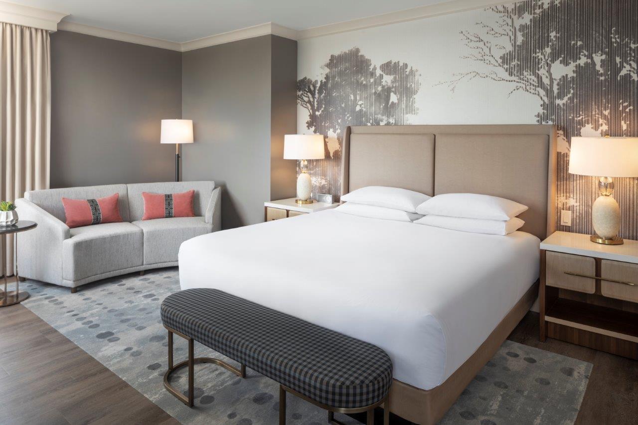 Upright bedroom with bed, sofa, lamps, and wall mural of trees, neutral and soft colors, wooden floor, and patterned rug.