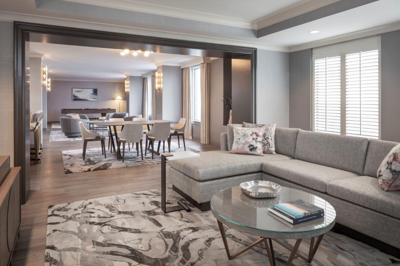 Living room with gray sectional sofa, round glass coffee table, and patterned rug, opening to a dining area with chairs and table.