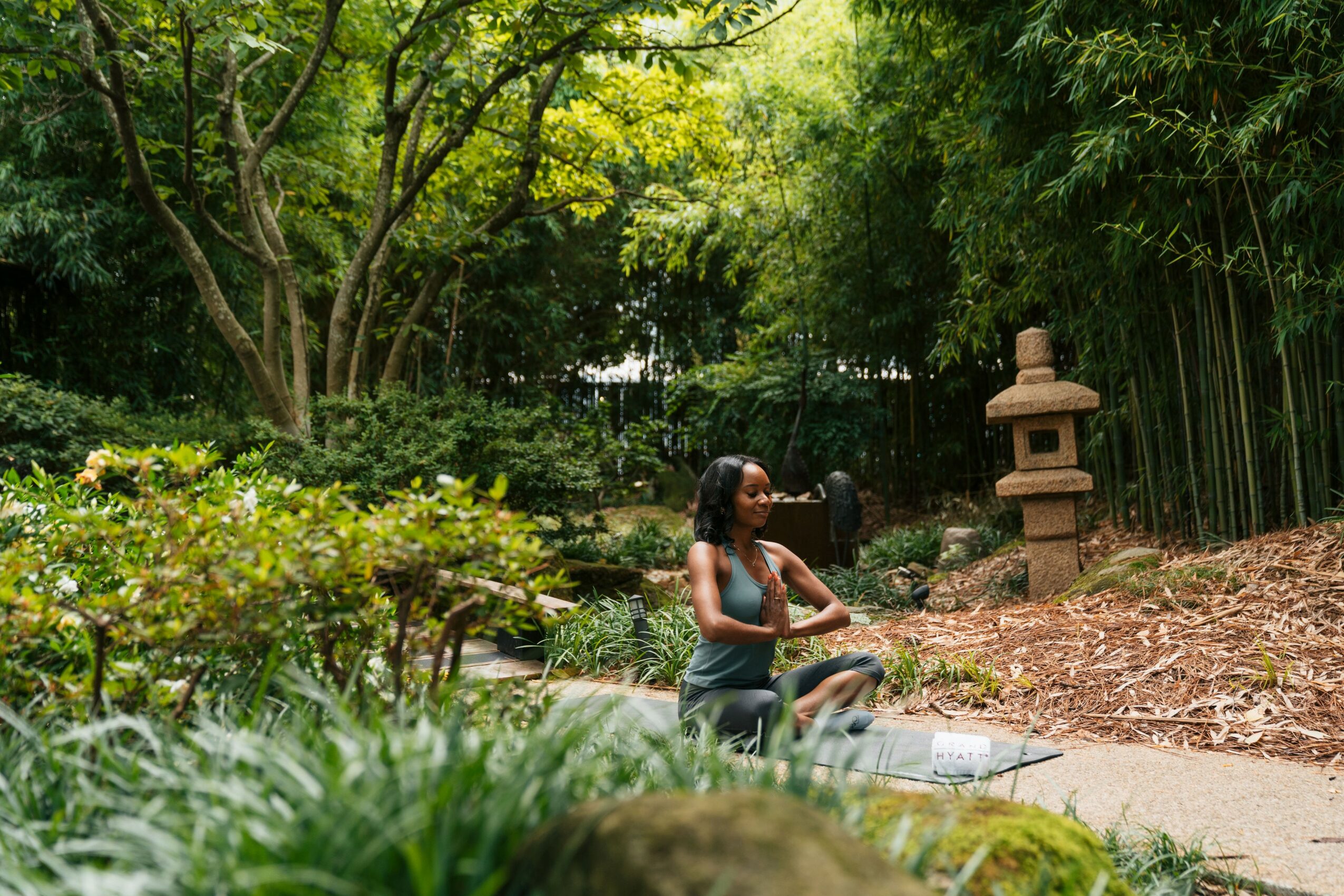 Person sitting on a stone in a lush garden with trees and a stone lantern, surrounded by greenery.