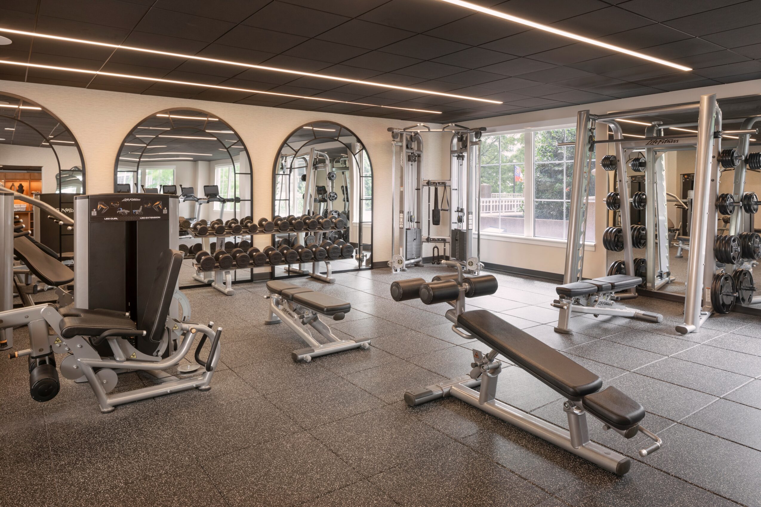 Gym with weight benches, dumbbell racks, and exercise machines, illuminated by natural light through large windows.