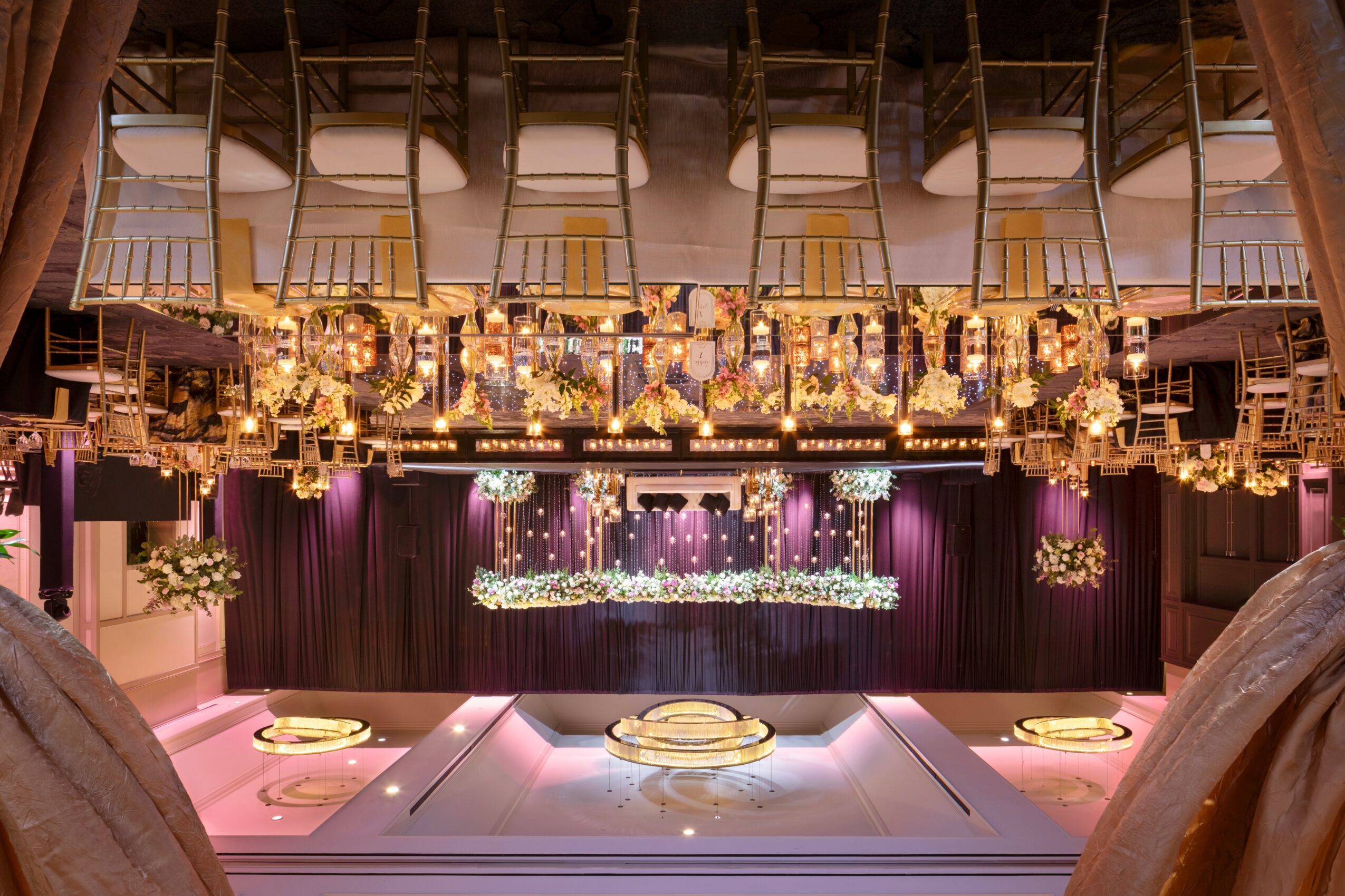 Upside-down view of a stage with purple curtains, decorated with hanging lights and floral arrangements, and a seating area with tables and chairs.