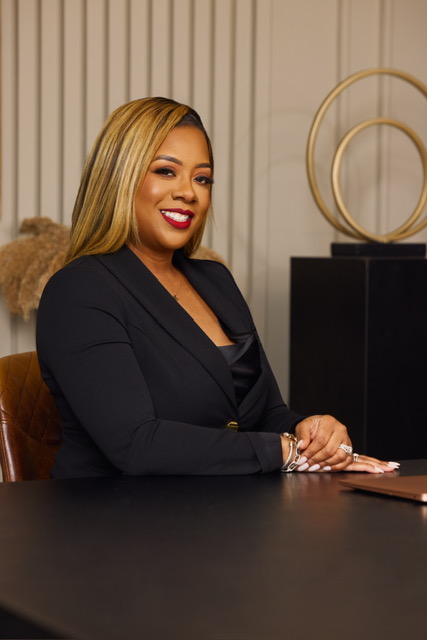 Woman with blonde hair and red lipstick sitting at a desk, smiling, in a professional setting.