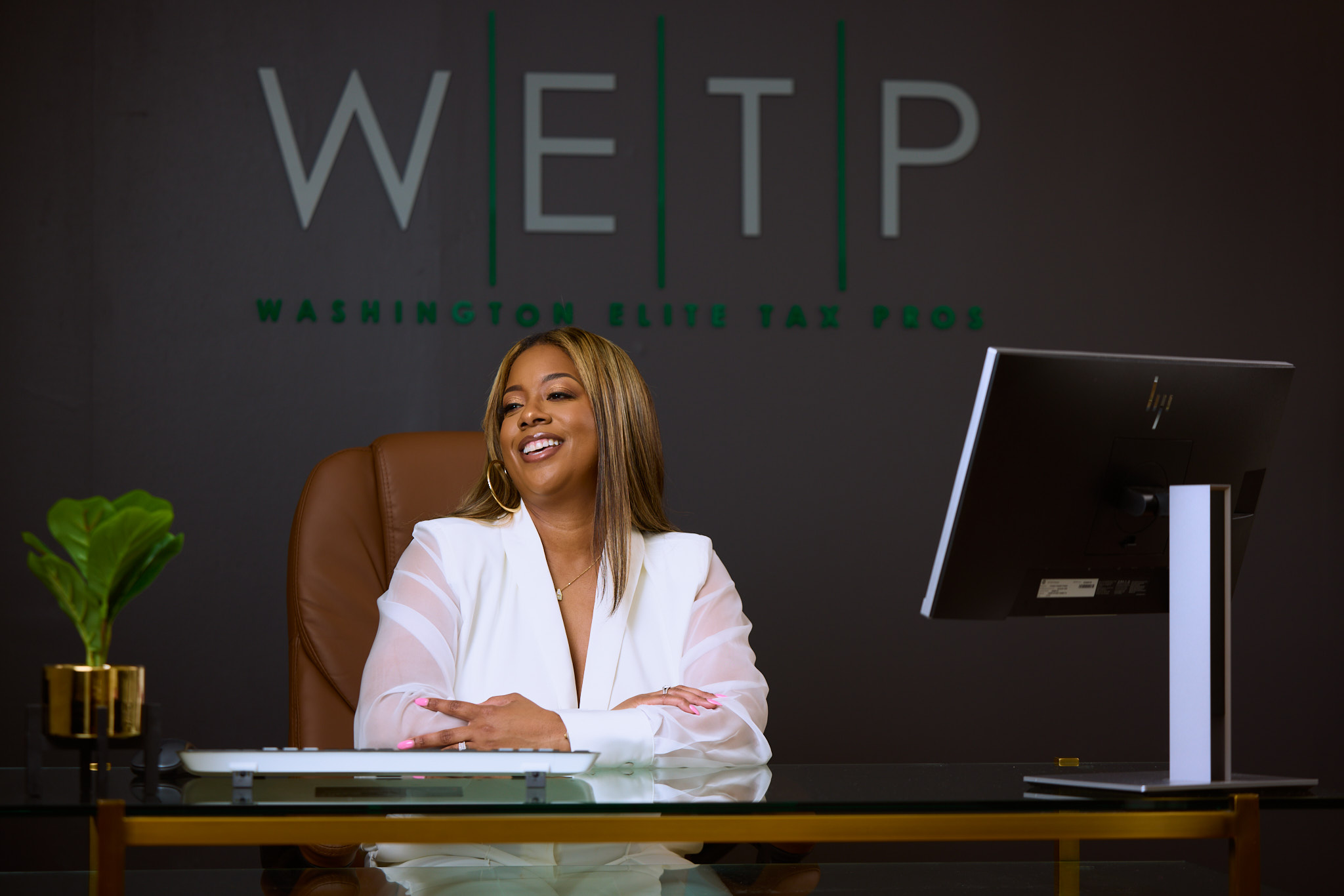 Woman sitting at desk smiling, with a computer monitor and a potted plant, in front of a dark wall with WETIP logo.