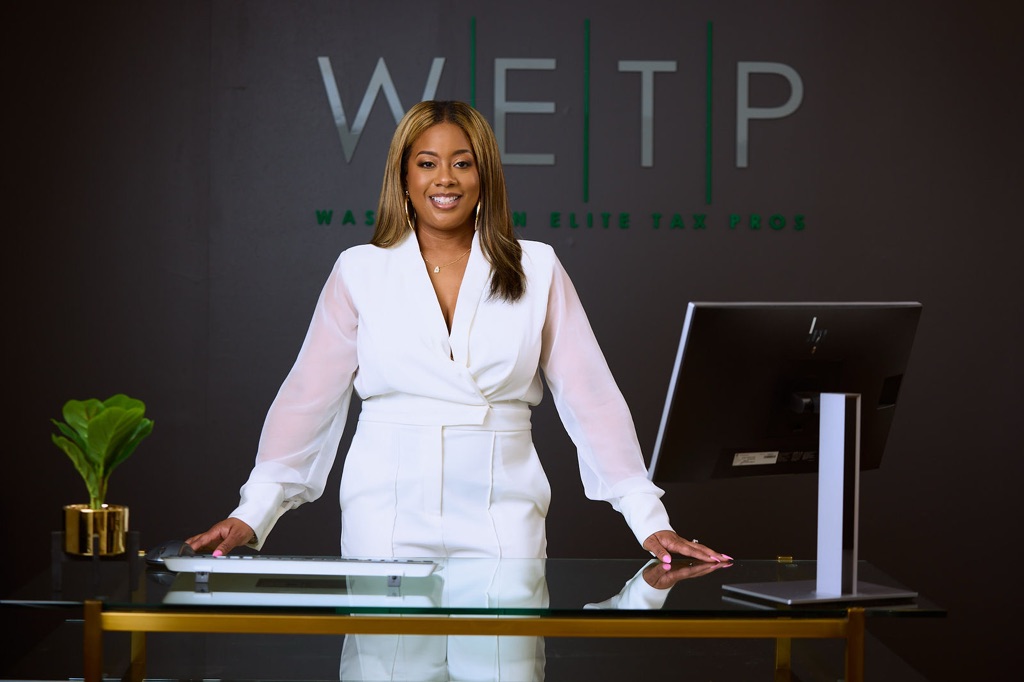 Woman standing behind a desk with a computer and a plant, smiling, in front of a dark background with text.