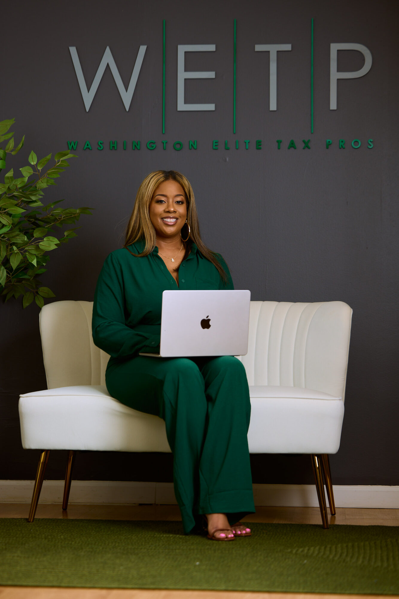 Woman sitting on a white sofa holding a laptop, smiling, in front of a dark wall with text, green plant nearby.