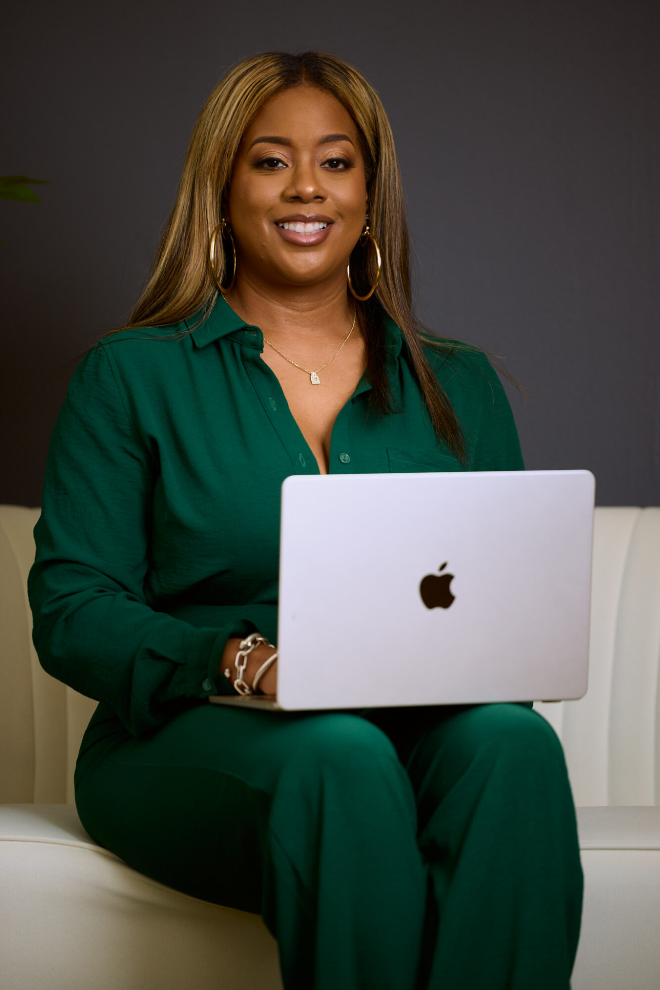 Woman with long hair in green outfit sitting with a laptop, smiling, dark background.