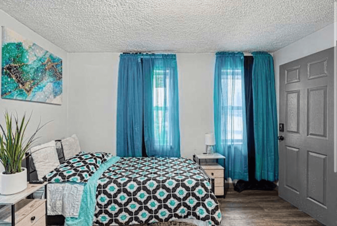 Bedroom with two windows, blue curtains, bed with patterned quilt, nightstand, and potted plant, gray door on the right.