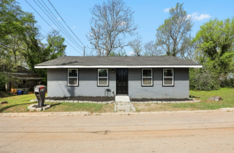 Single-story house with gray exterior, four windows, black front door, small front steps, and trees in background.