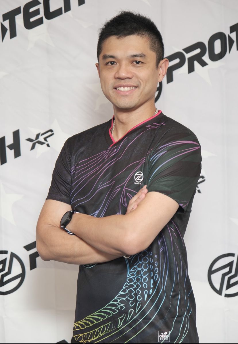 Young man with short black hair smiling, arms crossed, wearing a black sports jersey with colorful patterns, standing in front of a branded backdrop.
