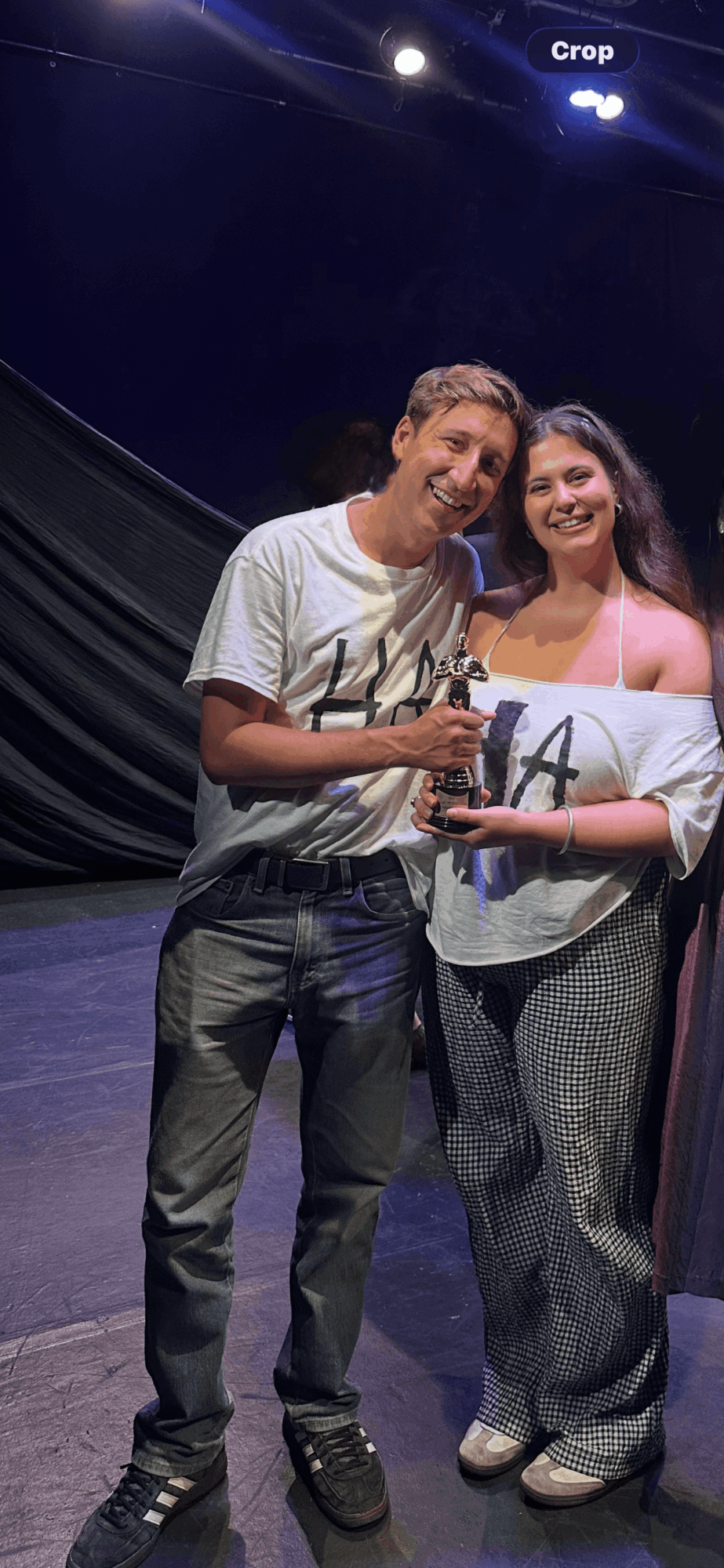 Two people smiling and holding a certificate or award on stage, with a dark background and stage lighting.