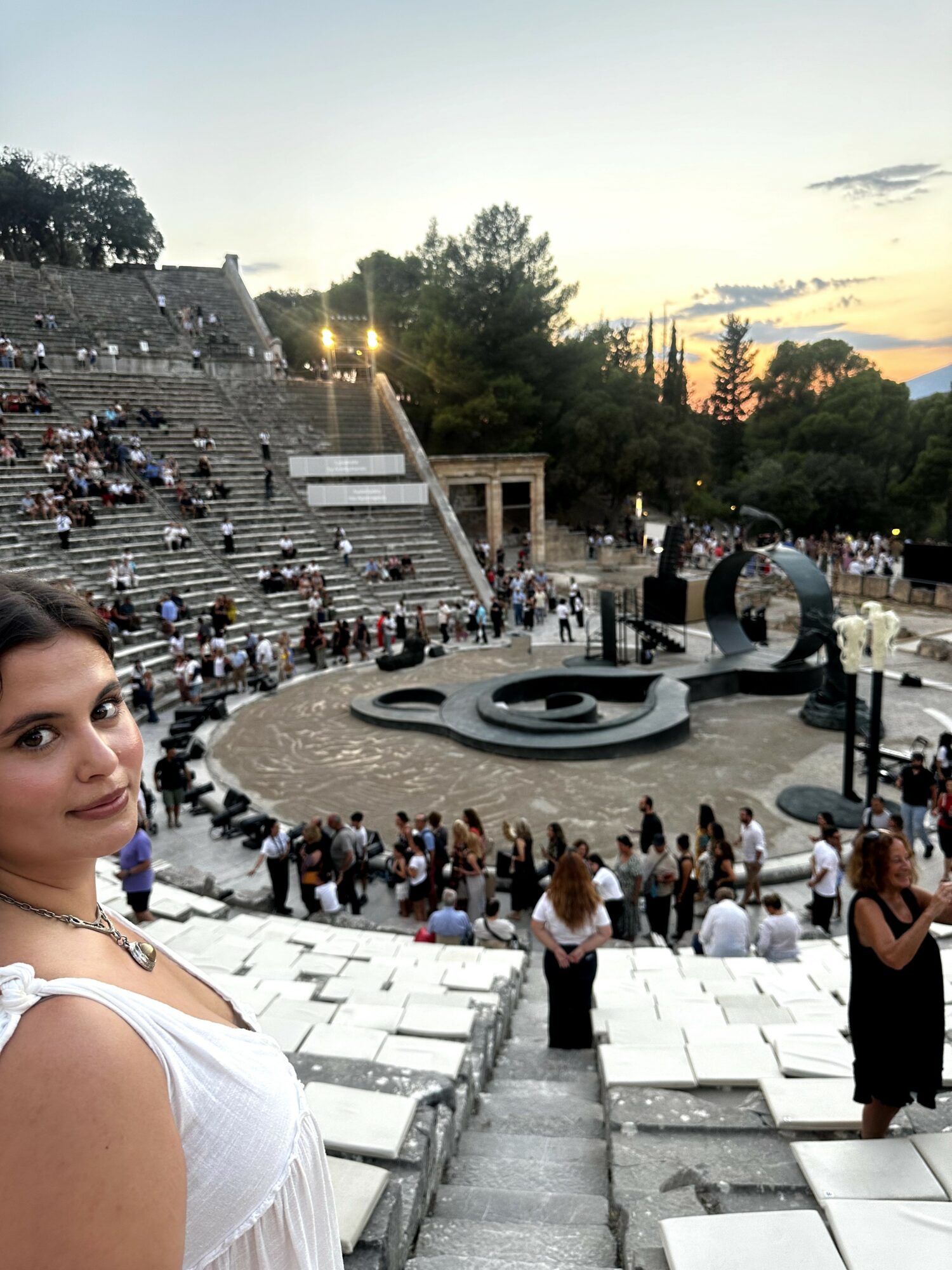 Woman in white dress at ancient amphitheater during sunset, with audience seated and standing around stage area.