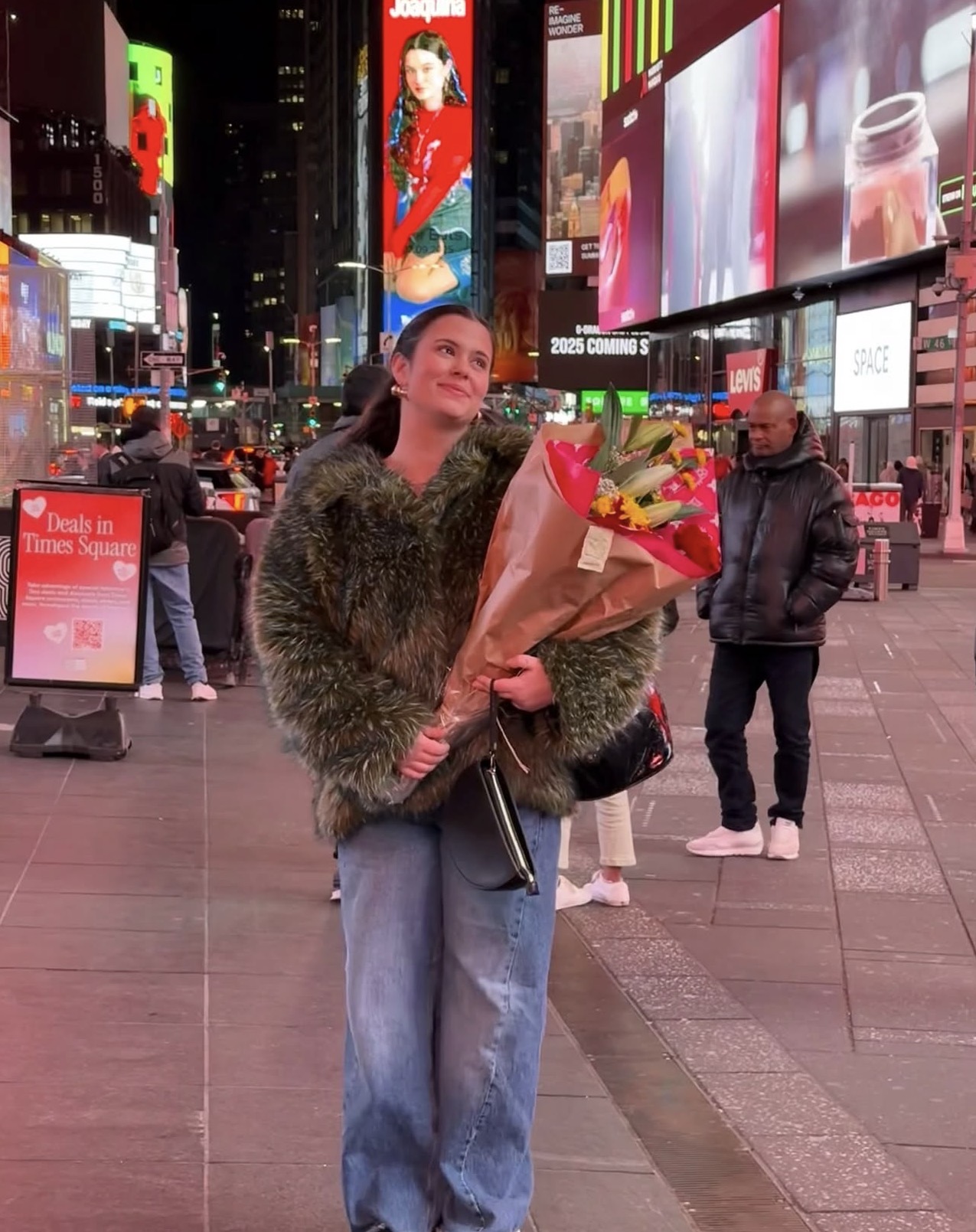 Woman holding a bouquet of flowers in a busy city square at night, illuminated billboards in the background.
