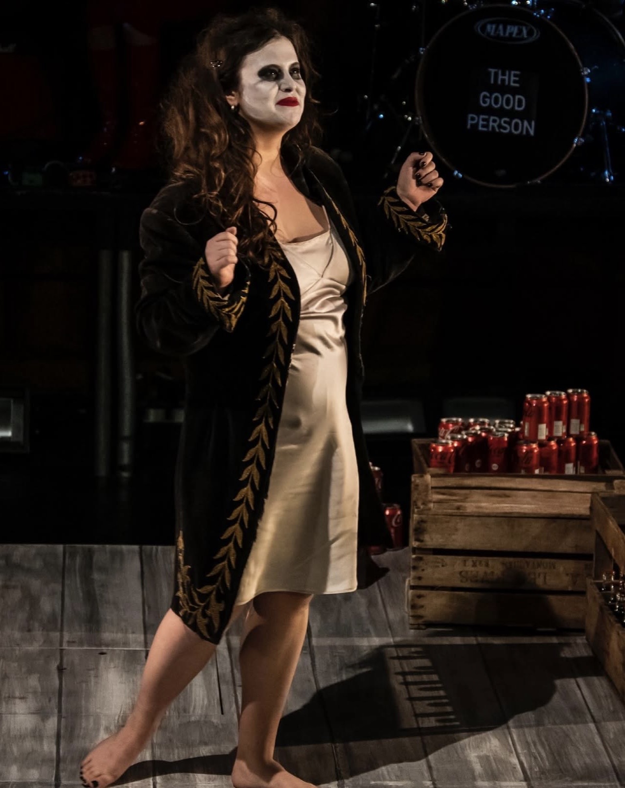 Woman with dark hair and makeup standing indoors, wearing a beige dress and black coat with gold details, next to a crate of bottles.