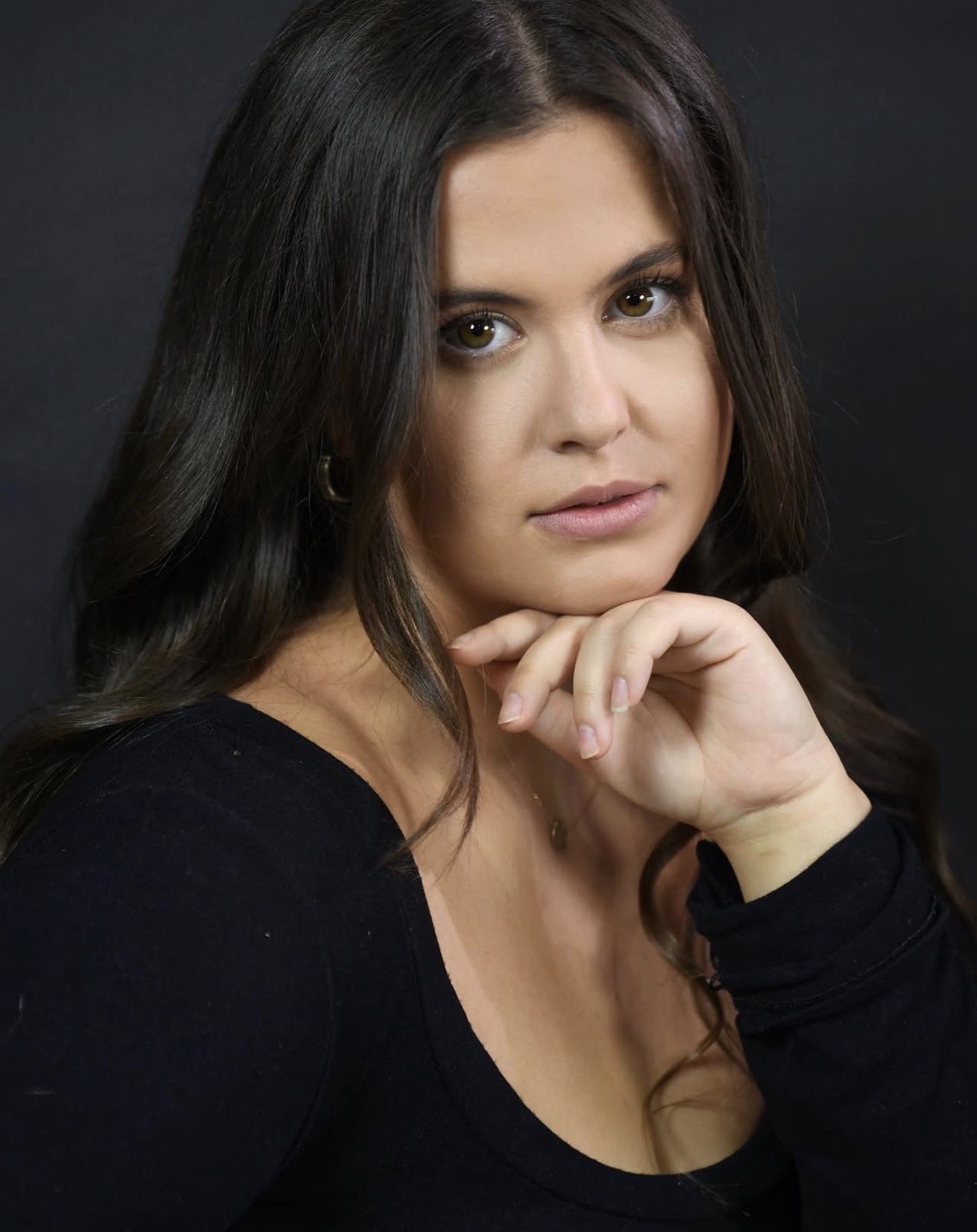 Woman with long dark hair and light skin, resting her chin on her hand, looking at the camera.