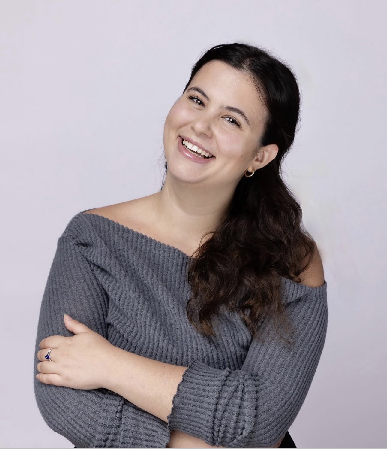 Young woman with long dark hair smiling, wearing a gray off-shoulder sweater, arms crossed, against a plain light background.