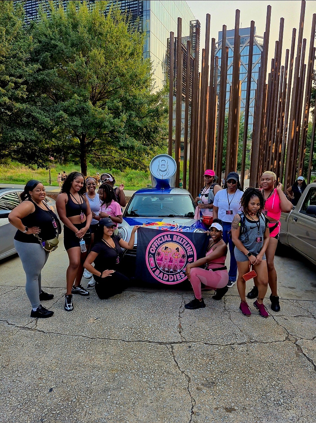 Group of women and girls posing in front of a police car with a pink logo, outdoors with trees and tall wooden poles.