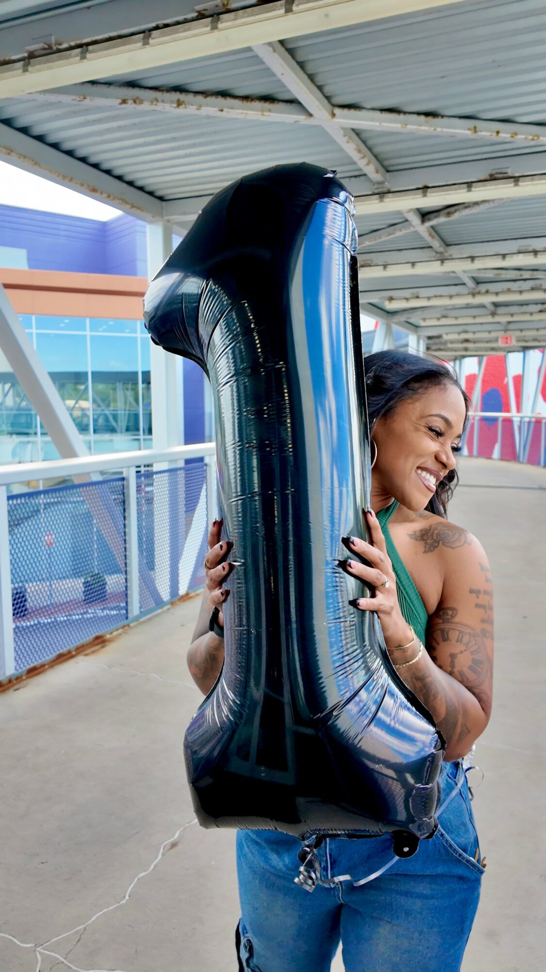 Woman smiling and hugging a large black number one balloon outdoors under a metal roof.