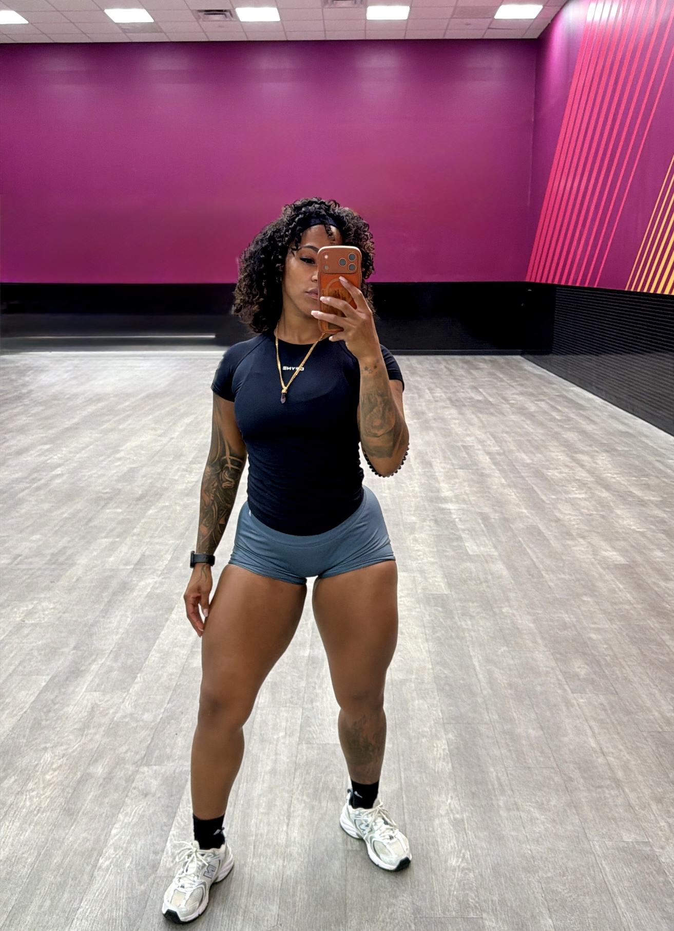 Person taking a mirror selfie in a gym with purple and pink walls, wearing a black shirt, blue shorts, and white sneakers.