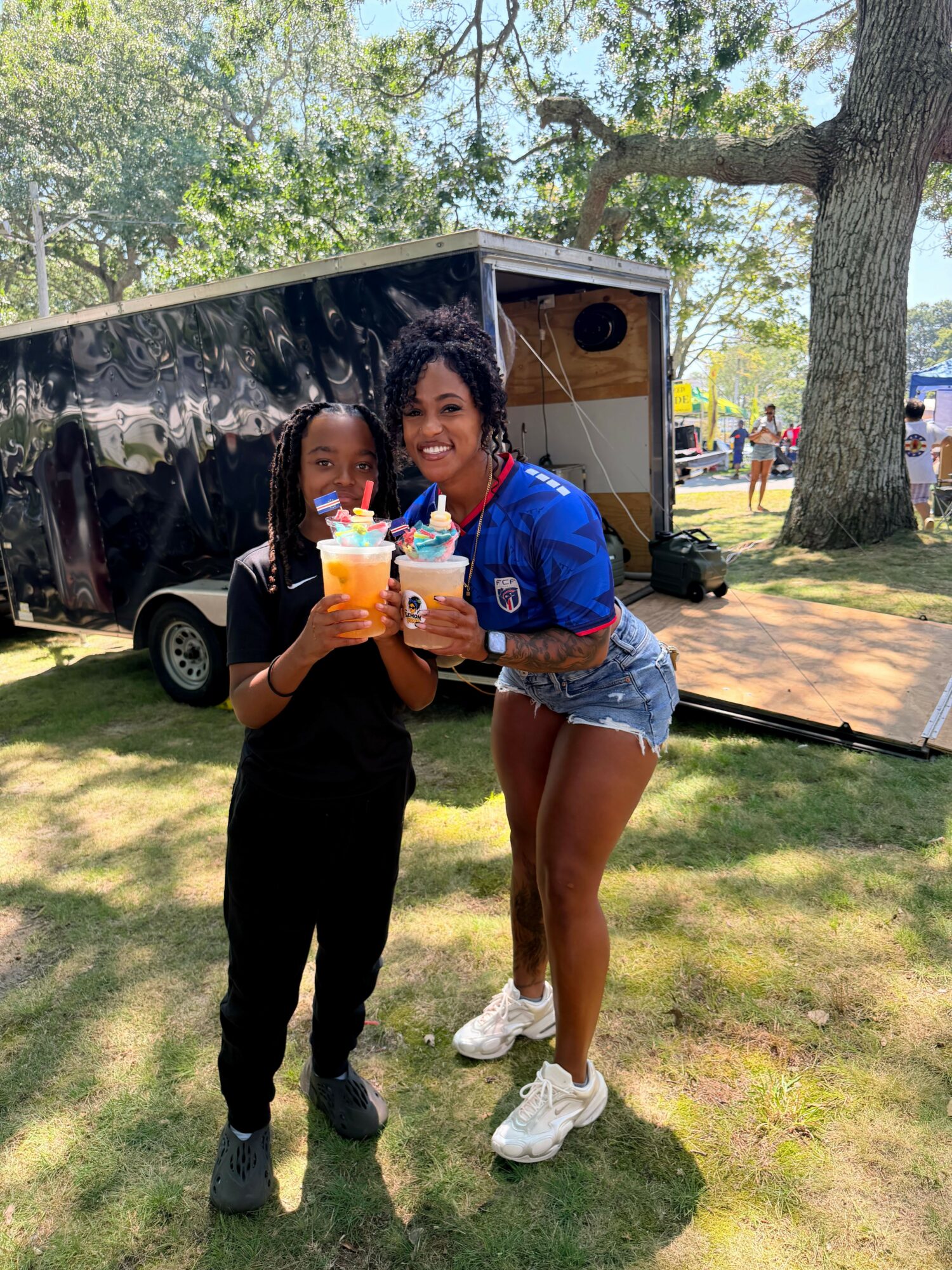 Two women standing outdoors on grass, one holding a drink, with trees and a trailer in the background.