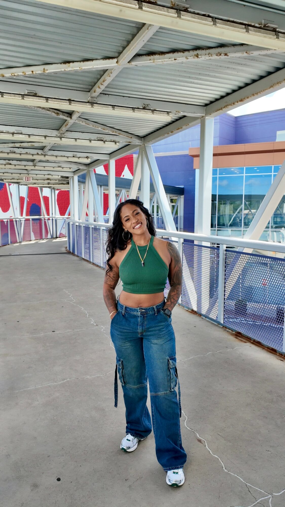 Young woman with long dark hair in a green crop top and baggy jeans standing on a walkway with a modern building in the background.