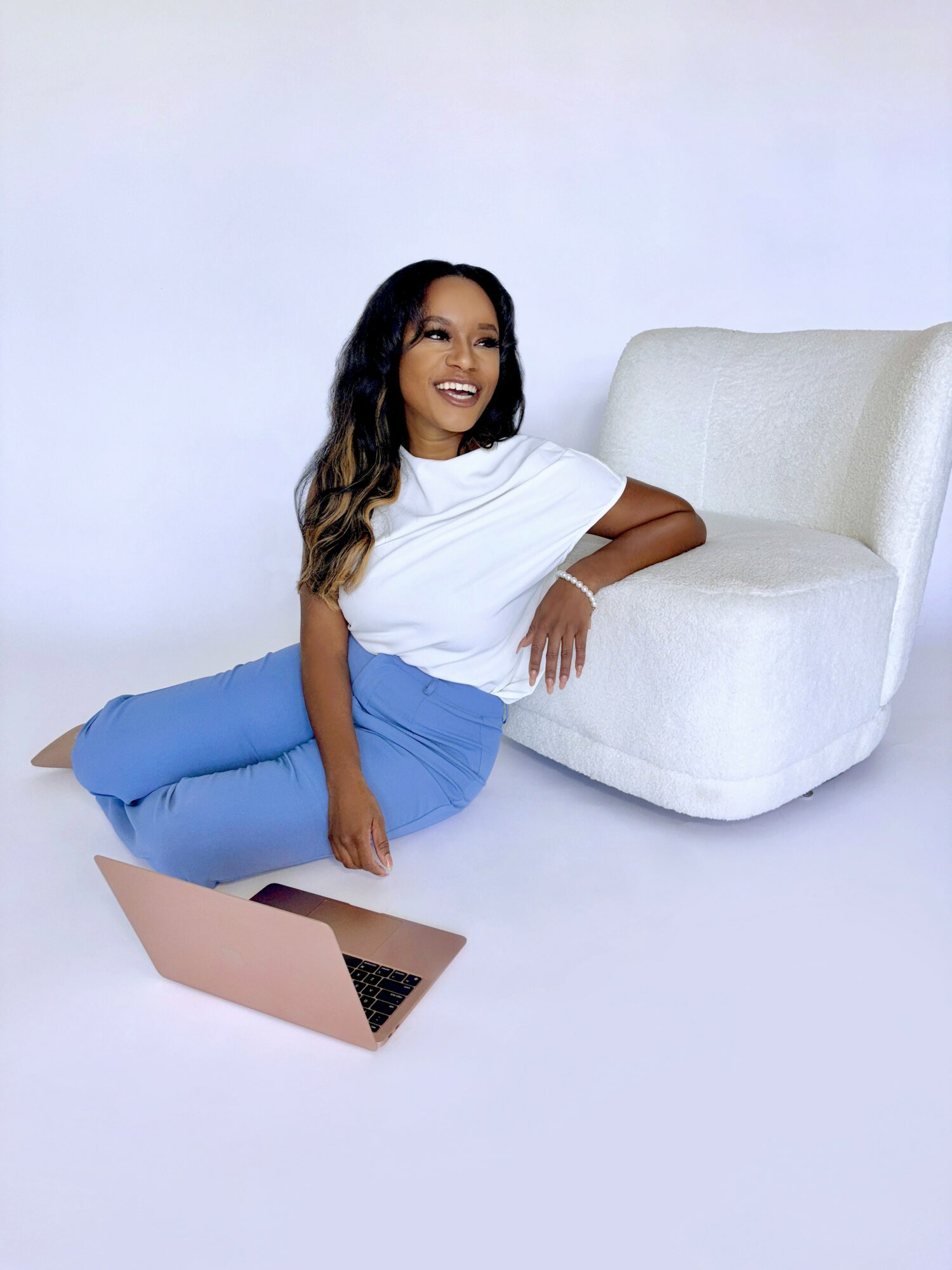 Woman sitting on the floor, leaning against a white armchair, smiling, with a laptop nearby, against a plain background.
