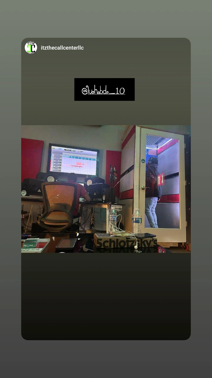Recording studio with computer, sound equipment, and a person standing near a glass window, viewed from a desk.