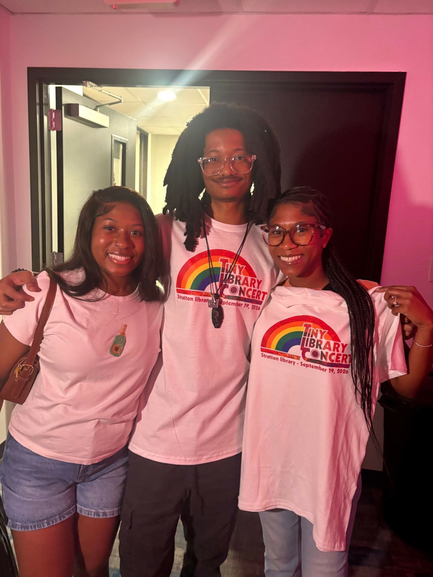 Three women standing together indoors, smiling, wearing matching pink T-shirts with rainbow designs, in front of a dark doorway.